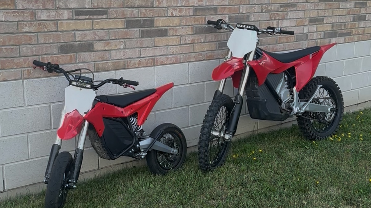 Two red and black electric dirt bikes parked side-by-side on grass against a brick and concrete block wall.